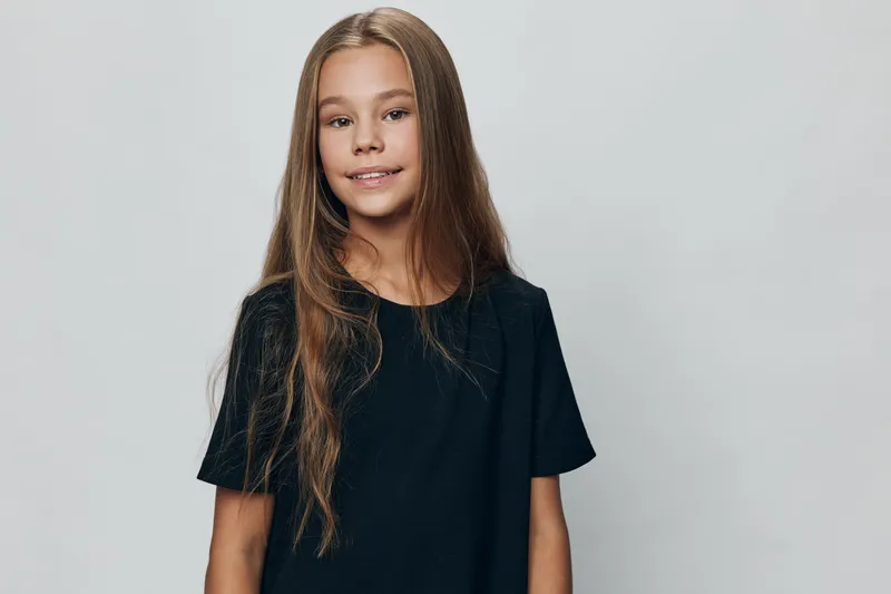 Portrait of confident girl with long hair wearing black shirt standing against gray background. Smiling child looking at camera with relaxed expression and natural pose.