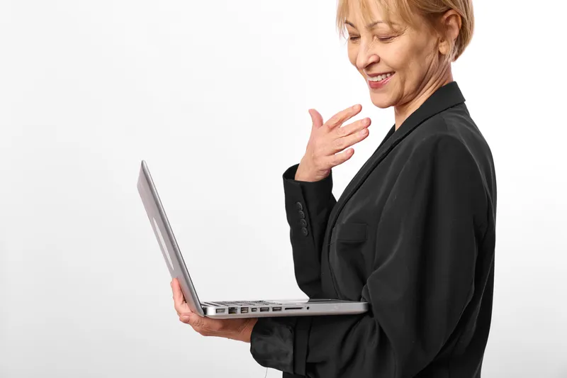 Elderly woman with laptop touches her neck, showing throat pain and discomfort, smiling softly, studio white background, concept of health concerns, stress, and cold symptoms