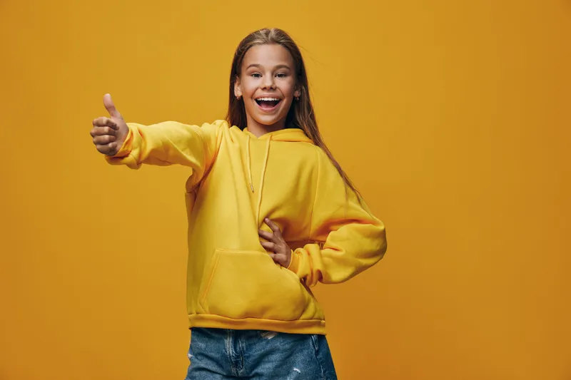 Girl with long hair smiling and showing thumbs up in yellow hoodie standing on orange background in studio for positive expression and happiness concept.