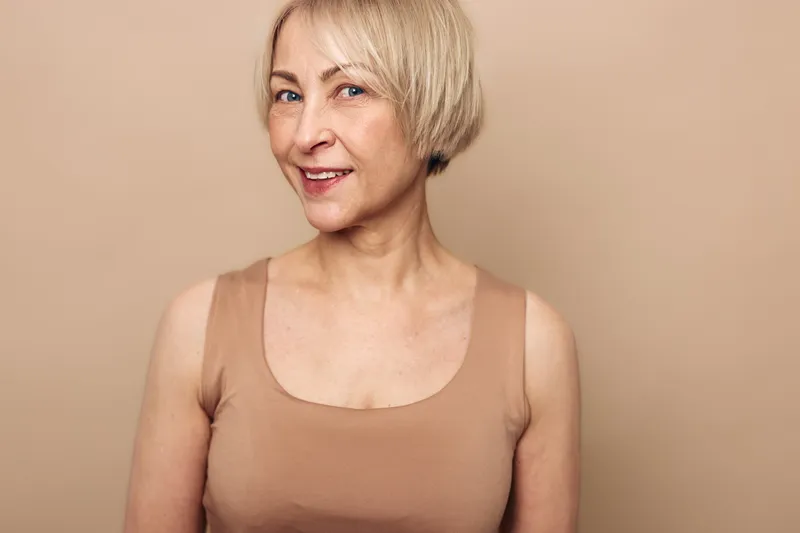 Smiling middle aged woman with short blonde hair wearing beige tank top stands against plain background. Concept for confidence, natural beauty, and mature lifestyle portrait.