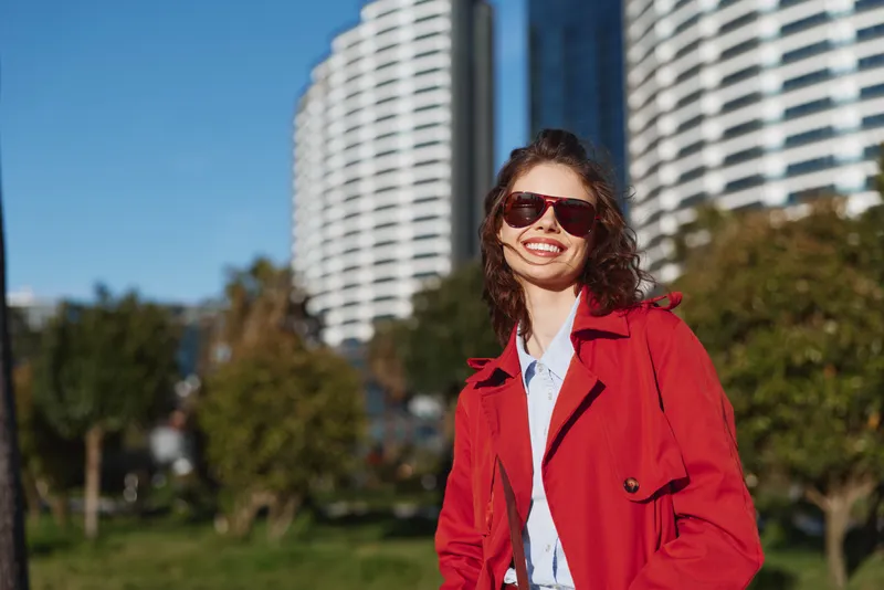 Young woman with curly hair smiles confidently wearing a red coat and sunglasses outdoors in an urban park on a sunny day.