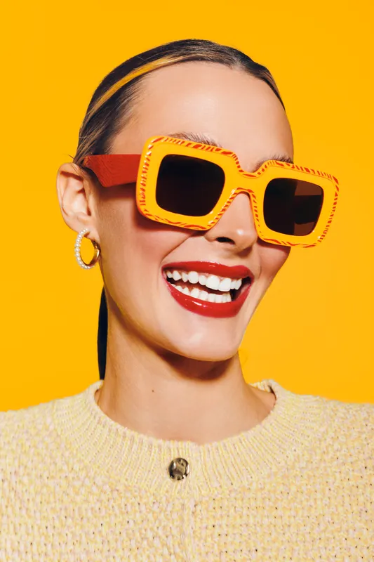 Smiling woman with bright red lipstick wearing large orange sunglasses and yellow sweater on vibrant yellow background. Concept of fashion, summer style, and joyful mood.