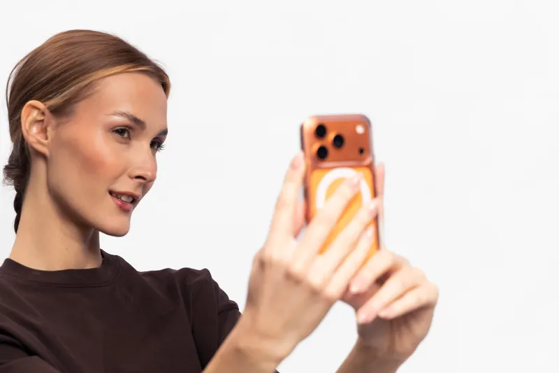 Young woman with light brown hair wearing dark shirt takes selfie with smartphone on white background. Concept of social media, communication, and modern technology usage.