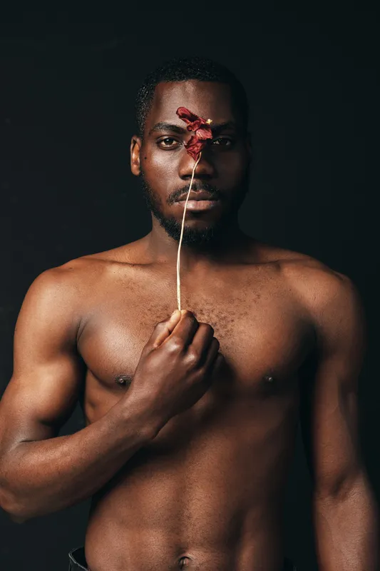 African man with dark skin and muscular body holding dried flower in front of face in creative conceptual visual style on black background. Artistic portrait with bare chest and serious expression.