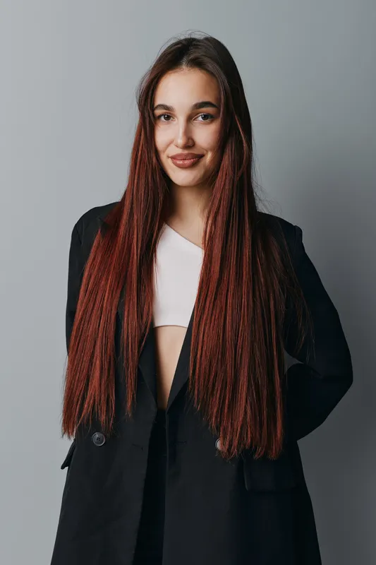 Young woman with long straight hair wearing black blazer and white top standing against gray background looking at camera with confident smile and relaxed posture in studio.