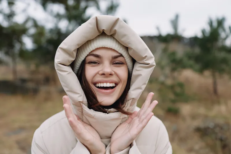 A cheerful woman outdoors in winter, smiling while wearing a warm coat with a hood. Cozy outerwear and natural light create a joyful candid portrait in cold scenery