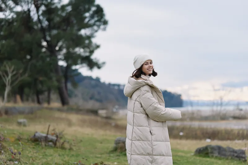 Woman in a light puffer coat and beanie stands outdoors by a lake, calm landscape, cool weather, contemplative mood under an open sky and gentle breeze.
