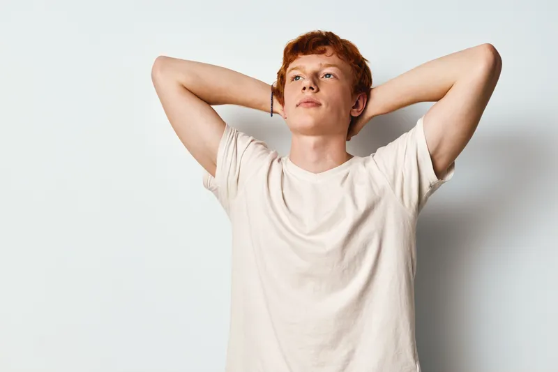 Young man with red hair wearing white shirt standing with hands behind head, looking thoughtful and confident against white background in casual style portrait.