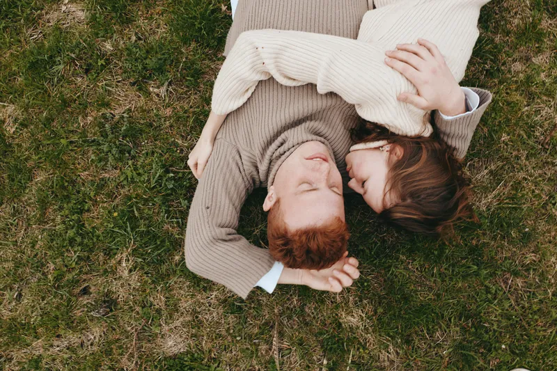 A couple lying on grass, facing each other, sharing a quiet moment outdoors. Cozy sweaters create a warm mood. Natural daylight highlights tenderness, connection, and simple happiness in a relaxed