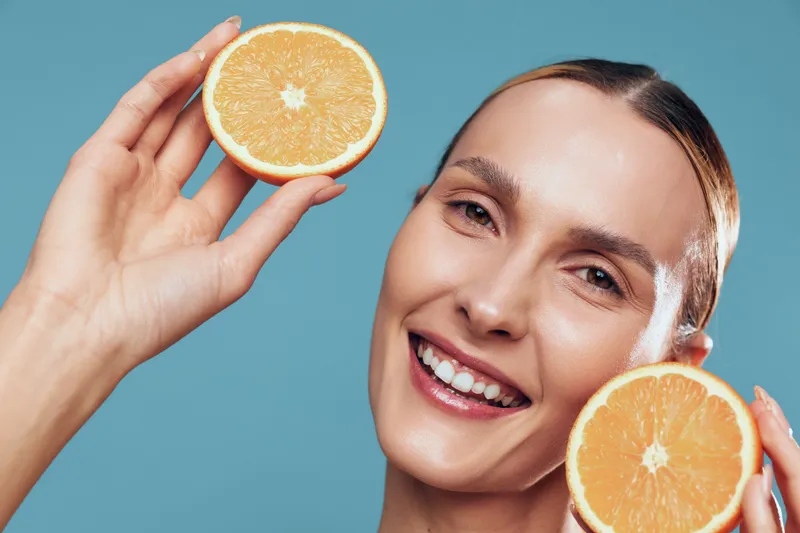 A smiling woman holds two halves of an orange close to her face against a blue background. The image conveys freshness, health, and natural beauty concepts.