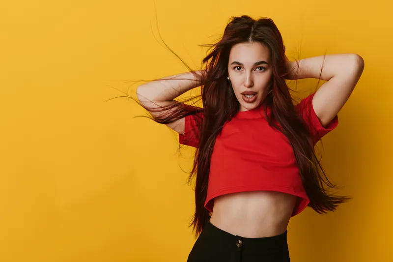 Young woman with long hair wearing red crop top and black pants posing with hands behind head against yellow background. Fashion, beauty and confident expression in studio shot.