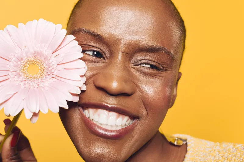 Close-up of happy African woman with dark skin holding pink flower near face on yellow background. Concept of beauty, natural smile, skincare, and floral wellness in studio lighting.