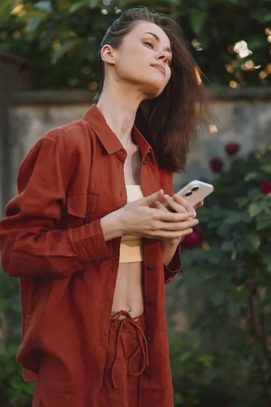 Stylish woman outdoors in a red jacket, holding a smartphone, casual yet confident, sunlight highlights her relaxed pose in a blooming garden, conveying modern lifestyle with natural colors