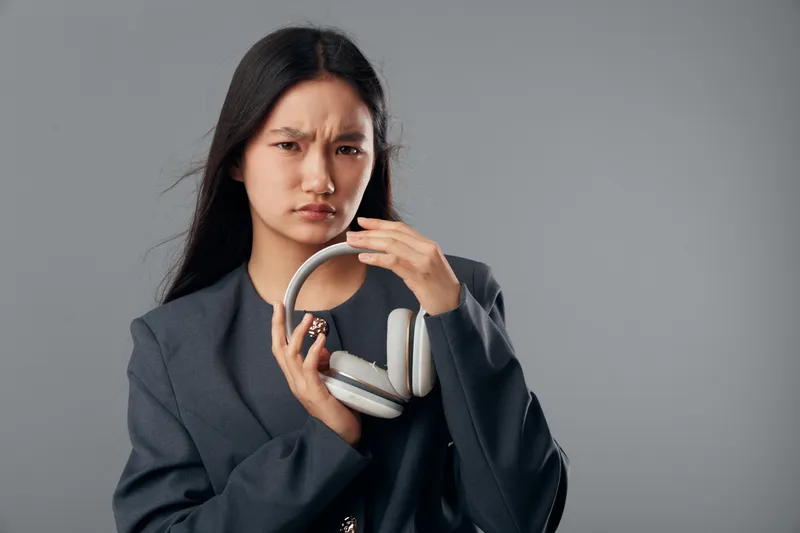 Young woman holding headphones with a confused expression against gray background. Female person in casual style showing doubt or discomfort with audio device in studio.