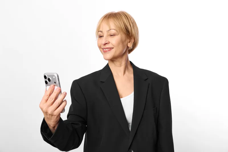 Smiling senior woman with smartphone, communication technology device, studio business portrait, holding phone near face, relaxed confident posture, bright neutral background, calm moment