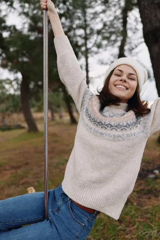 Woman on a swing outdoors, wearing a knitted sweater and beanie, smiling in a park setting, casual and playful moment among trees and grass.