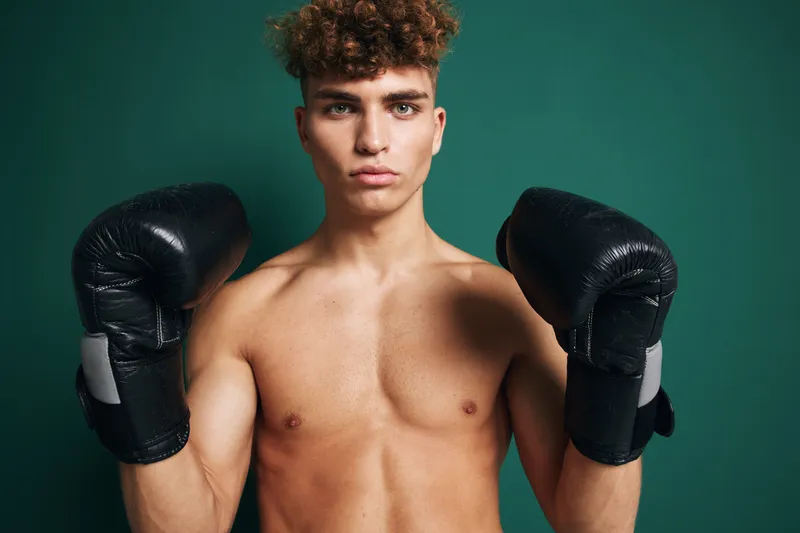 Young man with curly hair wearing black boxing gloves posing shirtless against green background for fitness, training, and combat sports concept with serious expression.