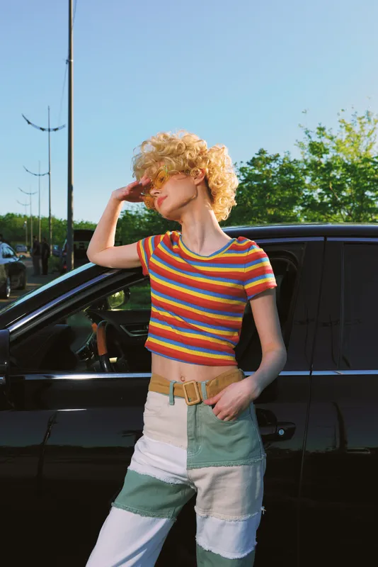 Cheerful woman outdoors, striped crop top, patchwork jeans, sunlit moment beside a car, relaxed pose, gentle breeze, vivid colors, nostalgic retro film mood, candid lifestyle