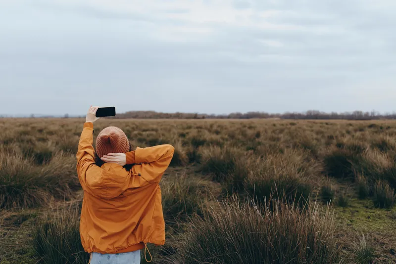 Woman stands in an outdoor field landscape, wearing an orange jacket and knit hat, holding a phone like a camera. Cloudy sky and grass create a nature lifestyle travel mood.