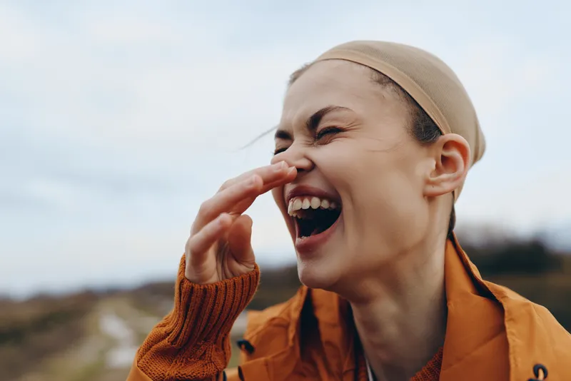 Woman lifestyle outdoors portrait of smiling woman in headscarf touching lips, orange jacket, natural light, open sky and countryside background, casual communication gesture