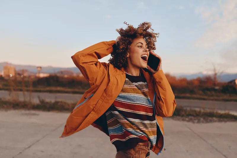 Smiling woman with curly hair outdoors, wearing an orange jacket and striped sweater, holding a phone to her ear while walking on a riverside path, warm sunset light, lifestyle moment
