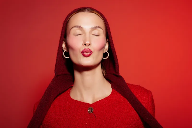 Young woman with closed eyes and red lips sends a kiss. She wears red clothes and a hood against a red background. Concept of love, affection, and beauty.
