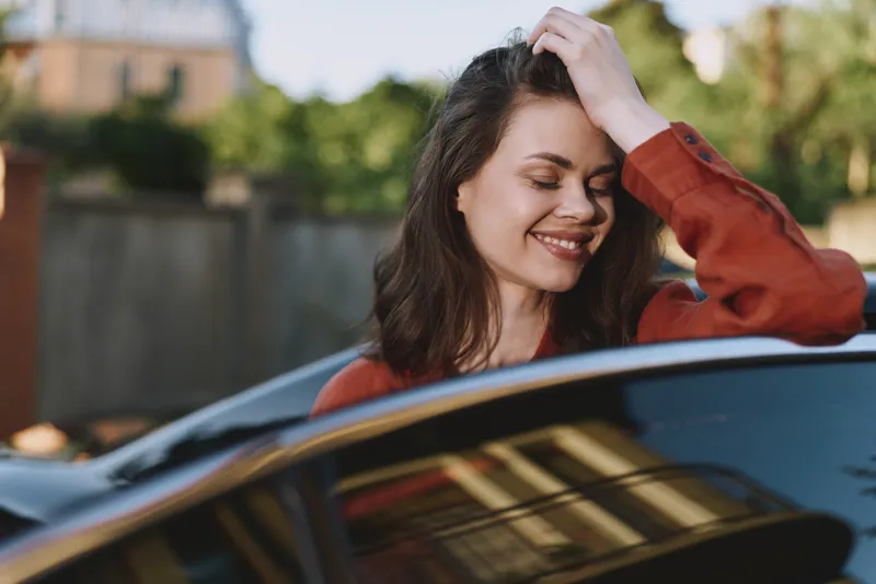 Casual woman enjoys a sunny moment by a car, smiling with her hand in her hair, relaxed lifestyle portrait highlighting everyday confidence and freedom
