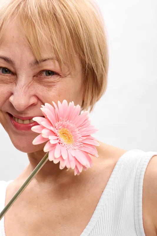 Closeup portrait of a blonde woman smiling with a pink gerbera flower near her cheek, soft light and gentle expression suggest calm happiness today