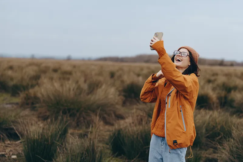 Woman lifestyle outdoors in grassy field, smiling with glasses, wearing orange jacket and knit beanie, arms raised, cloudy daylight, calm wind, natural colors, soft breeze, horizon