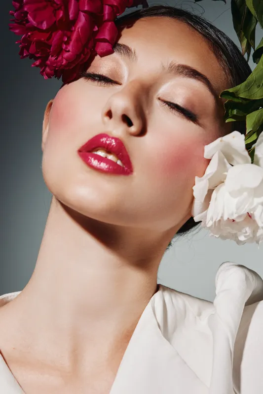 Close-up of a woman with glossy red lips and pink blush surrounded by red and white flowers. Soft lighting emphasizes her flawless skin in a creative beauty fashion concept.