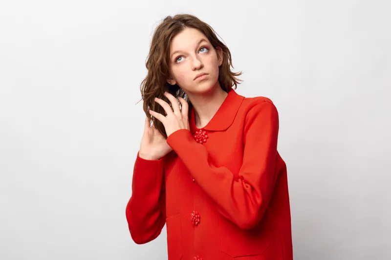 Young woman in red sweater adjusts her curly hair while looking up. Studio portrait on white background. Concept of casual style, fashion, and natural beauty.