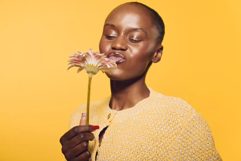 Portrait of a dark skinned African woman holding and smelling a pink flower on yellow background. Concept of natural beauty, floral, skincare, and positive mood in studio lighting.