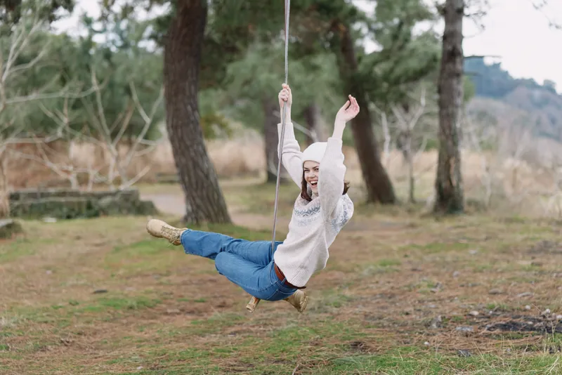 Woman on a swing in an outdoor park, carefree moment, smiling and enjoying fresh air, nature around, casual outfit, playful balance, rustic setting, serene afternoon, light breeze, candid lifestyle