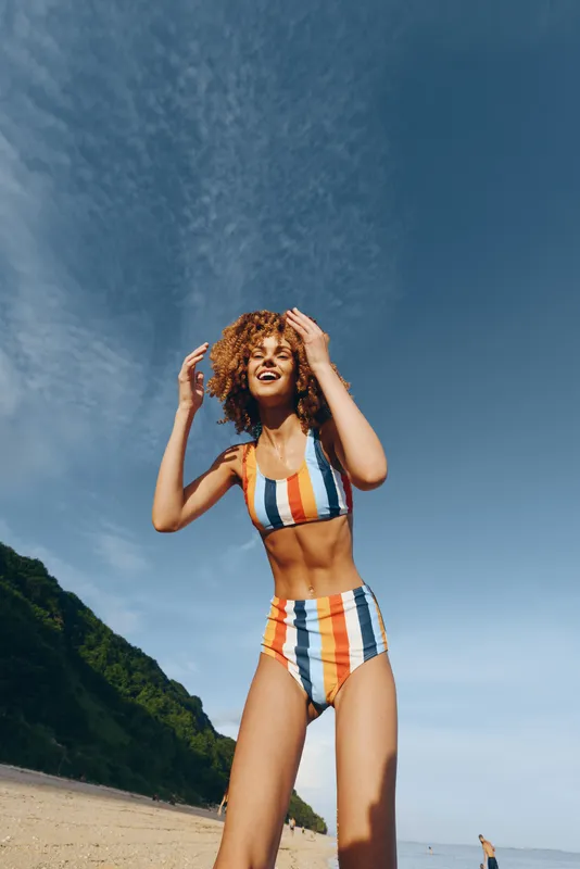 Woman in swimsuit on beach, summer outdoors, blue sky above mountains, smiling, hands on curly hair, outdoor portrait, sunny seaside landscape, vacation mood