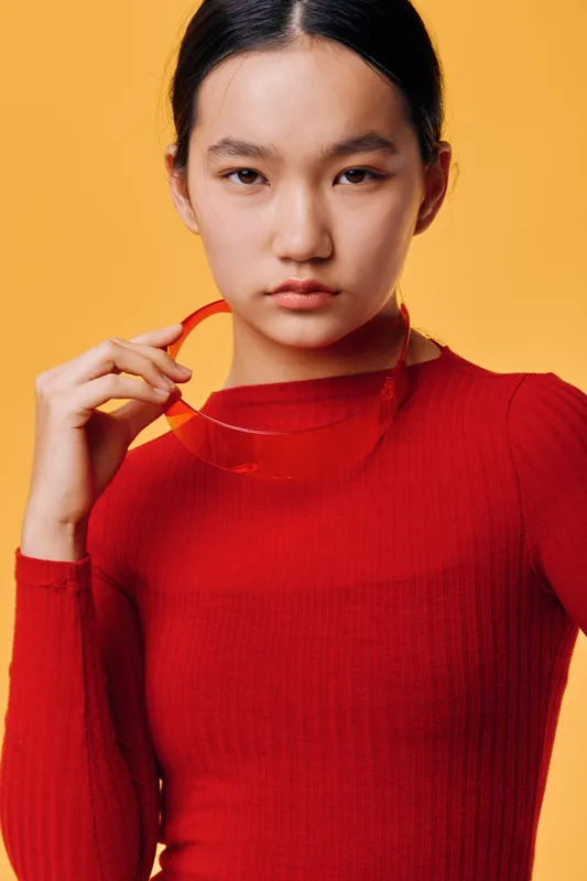 Young woman with serious expression wearing red sweater holding red sunglasses posing on yellow background. Asian female model with casual look and minimal makeup in studio portrait.