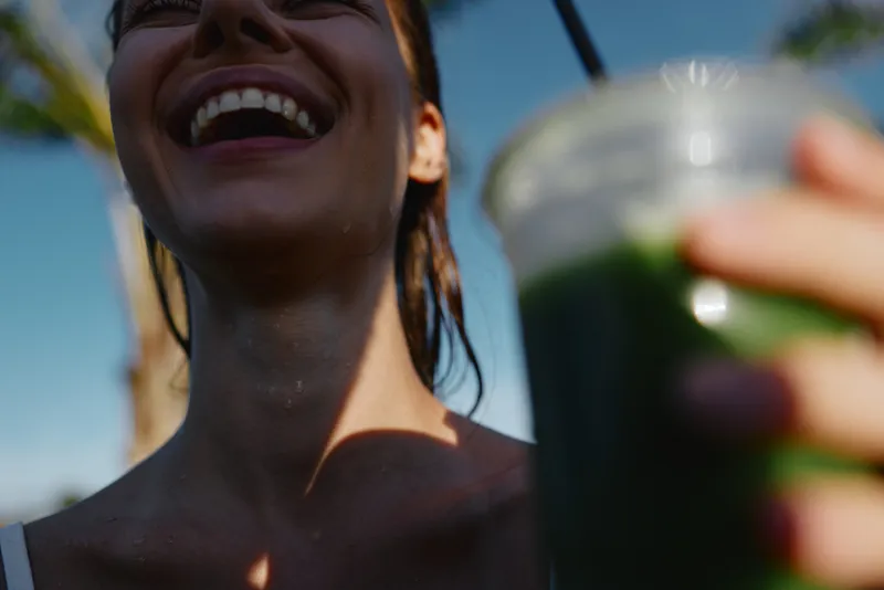 A woman outdoors smiles while a smoothie beverage is held close to the camera, capturing a sunny, candid moment of summer lifestyle, health, happiness, and refreshment.