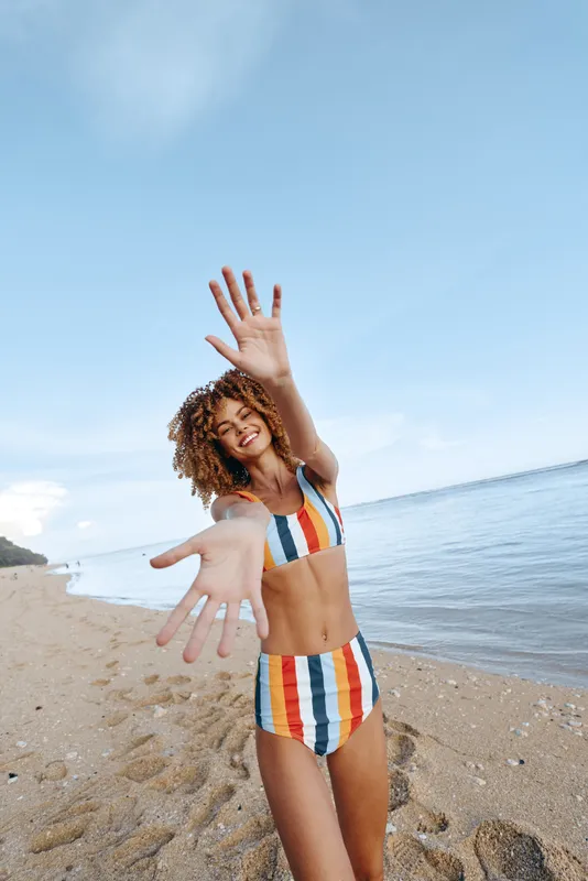 Happy young woman in colorful striped bikini smiling and reaching out to camera on sandy beach with ocean and clear sky background on sunny day.