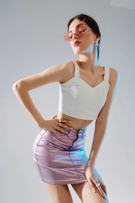 Young woman in a white top and shiny purple skirt poses confidently wearing pink sunglasses. Studio shot on a grey background. Concept of woman beauty fashion and style.