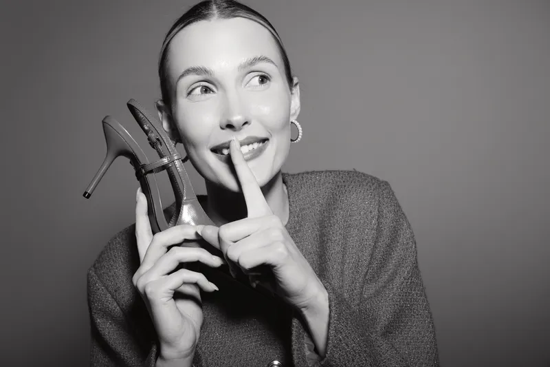 Young caucasian woman with light skin holds high heel shoes near face and makes shushing gesture, wearing earrings and jacket. Concept of fashion, style, and secrecy.