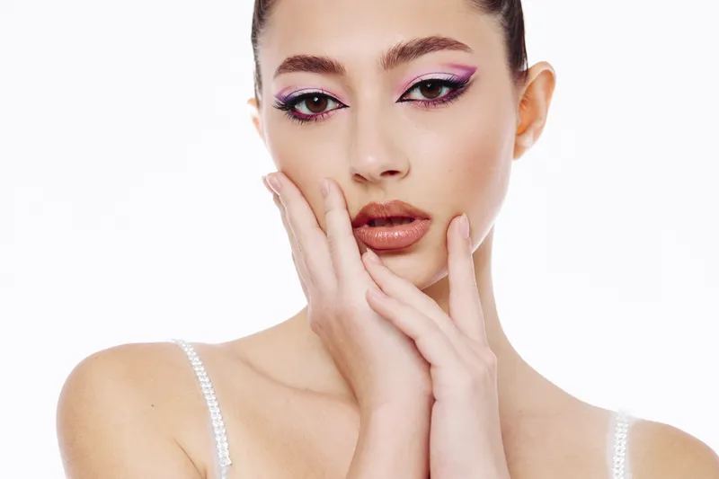 Closeup portrait of a young woman with creative purple eyeshadow makeup posing with hands near her face on a white background. Concept for beauty, fashion, and cosmetics.