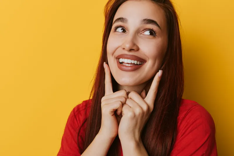 Young woman with long hair smiling and pointing fingers at her cheeks on yellow background. Happy female portrait with positive expression and casual red shirt in studio.