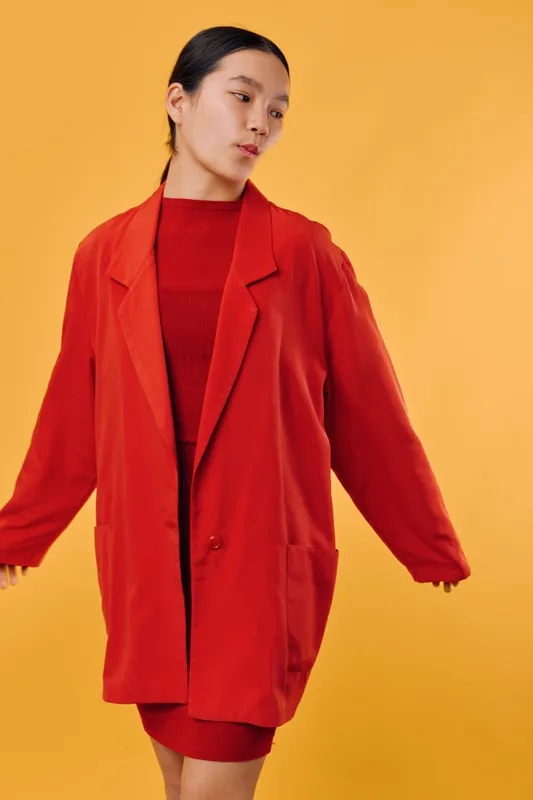 Young woman wearing oversized red blazer and dress posing against yellow background with minimal makeup and hair tied back. Fashion model looking sideways with casual confidence in studio.