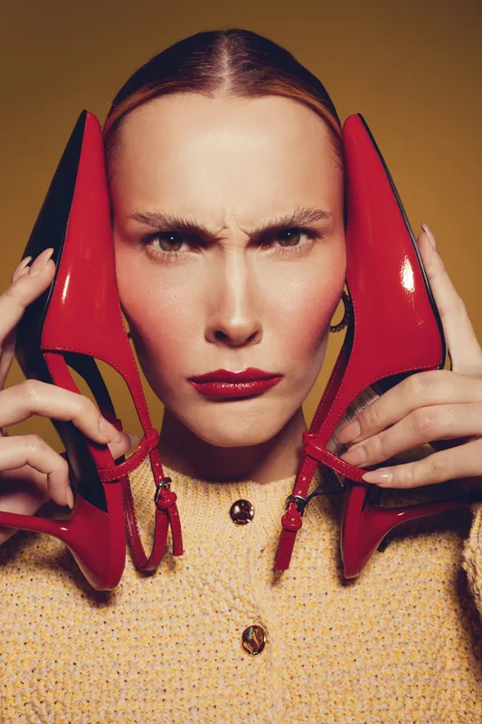 Close-up of serious woman holding red high heel shoes near face. Fashion concept with bold makeup and intense expression on yellow background. Stylish and modern portrait.