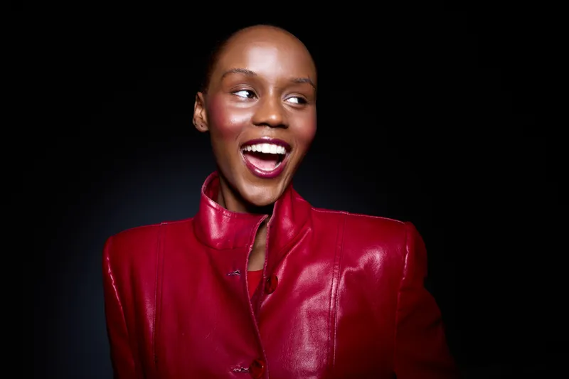 Smiling black woman with dark skin wears a red leather jacket, looking to the side with an open mouth. Studio portrait on black background. Concept of fashion, beauty, and confidence.