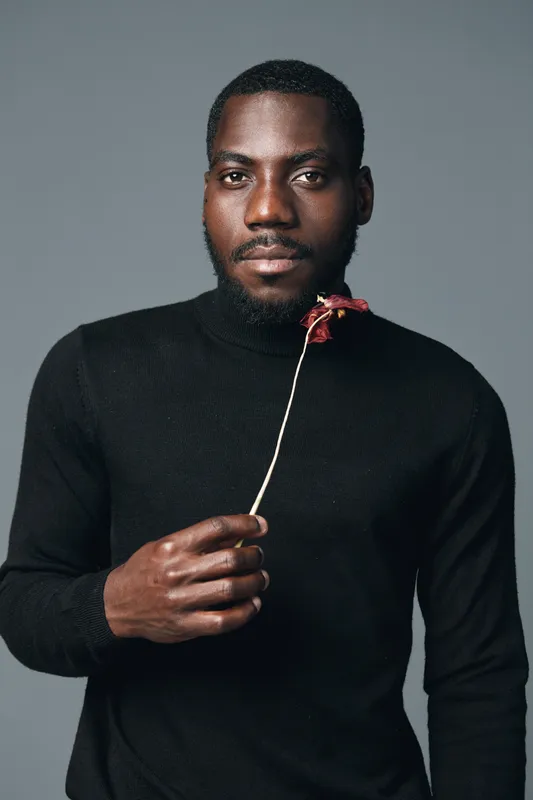 African man with dark skin wearing black turtleneck holding dried flower in creative conceptual visual style studio portrait with neutral background and serious expression.