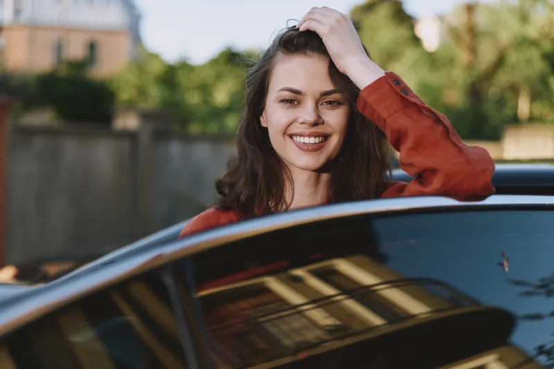 Woman smiling beside a car in an outdoor casual setting, showcasing lifestyle portrait with happiness, sunlight, and fashion elements in a relaxed street scene, hand in hair