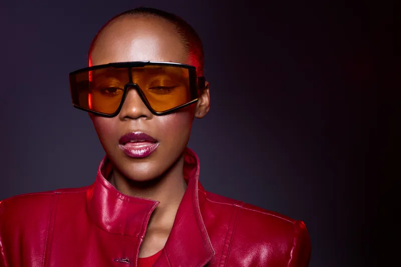 Close-up portrait of a confident black woman with dark skin wearing oversized orange sunglasses and a red leather jacket. Studio lighting highlights her glossy lips and smooth skin.