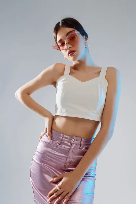 Young woman in fashionable pink metallic skirt and white crop top poses confidently wearing stylish pink sunglasses. Studio shot on a neutral background. Concept of woman beauty fashion.