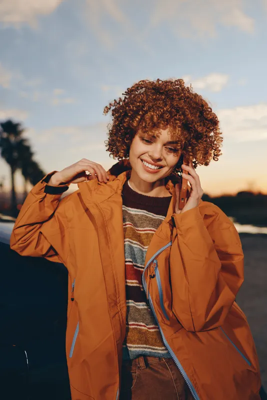 Woman smiling with curly hair in a jacket, portrait outdoors at sunset, warm sunlight and evening sky create relaxed lifestyle mood, beauty and selfcare in a casual outdoor moment