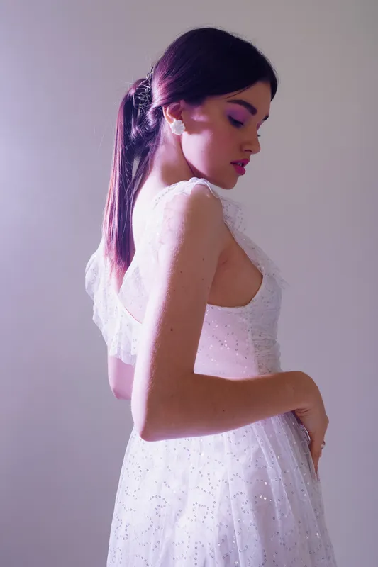 Young woman in a white dress poses sideways with closed eyes and pink makeup. Soft lighting highlights her beauty and fashion style in a minimal studio setting.